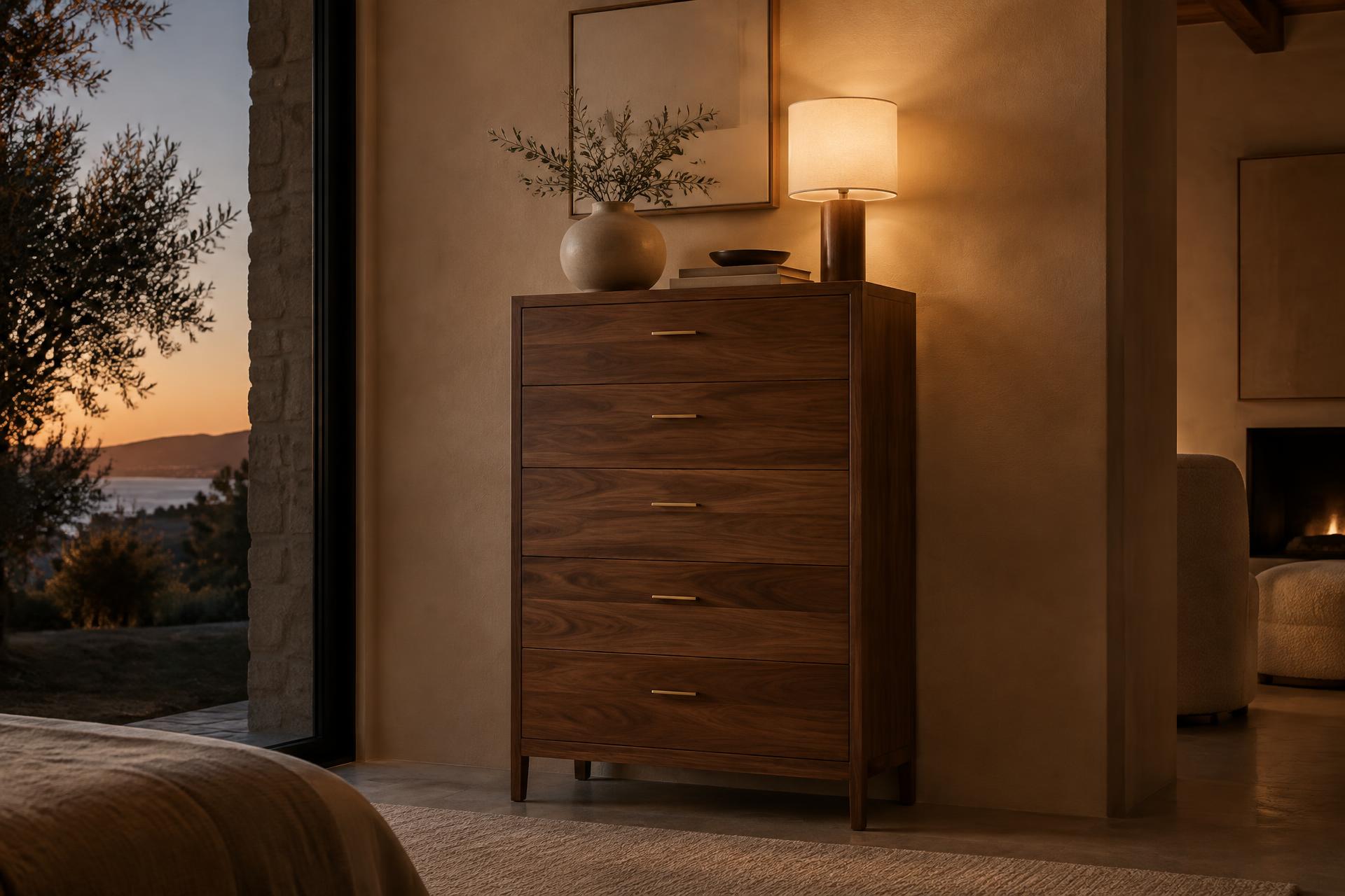 Tall walnut chest of drawers with brass pulls beside a lamp at dusk